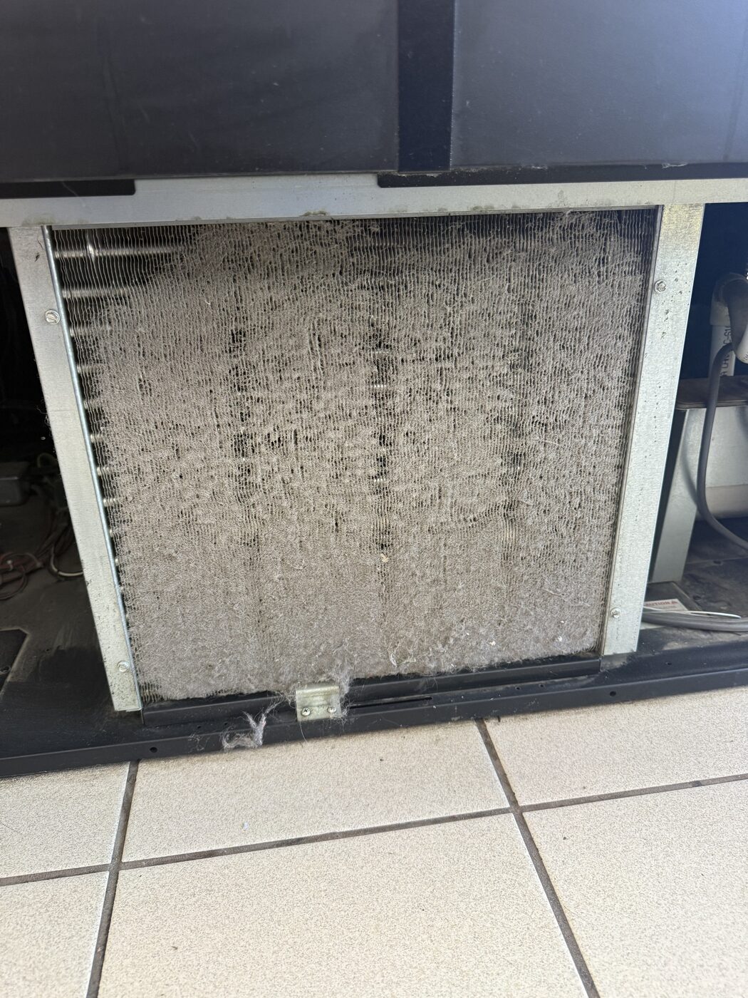 Heavily lint-clogged condenser coil under a commercial refrigerator on tile floor
