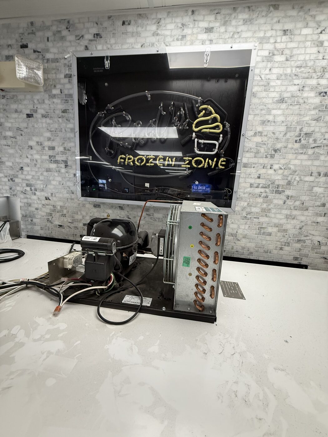 Compact refrigeration condensing unit with compressor and condenser coil on a counter below a Frozen Zone neon sign
